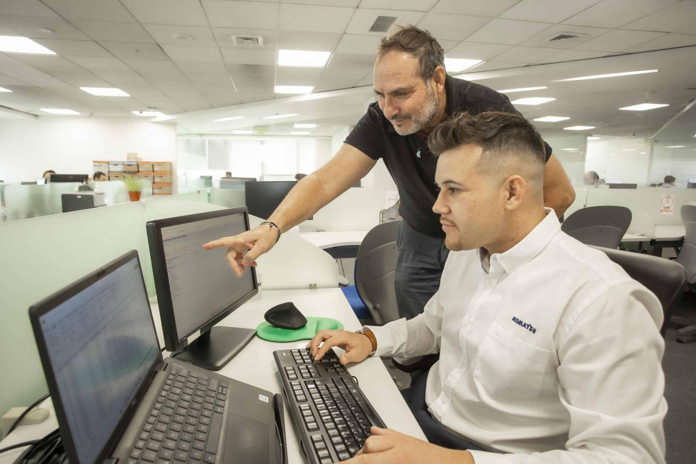 Man assisting colleague at dual monitors in modern office setting