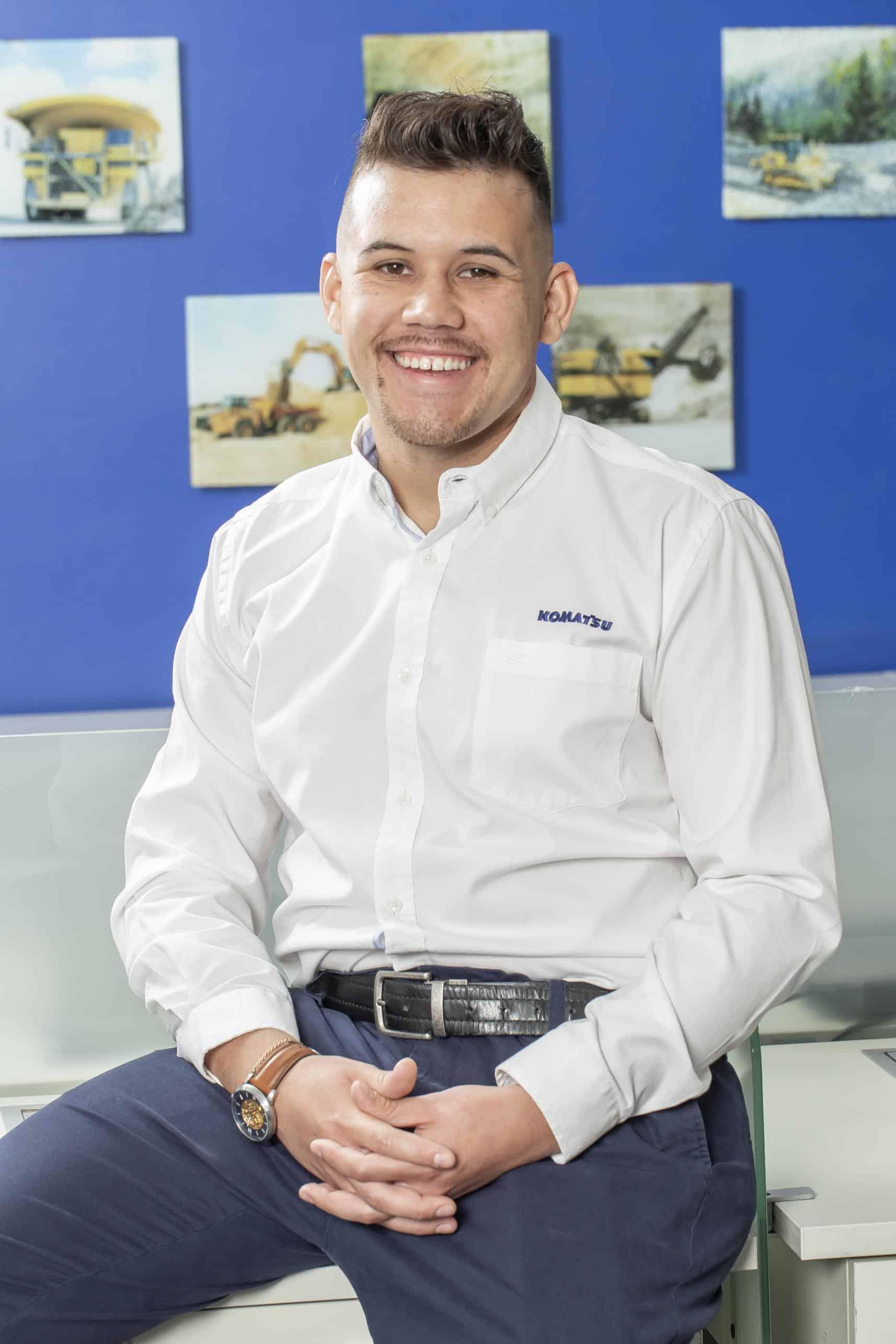Man smiling in Komatsu shirt seated in front of blue wall with machinery photos